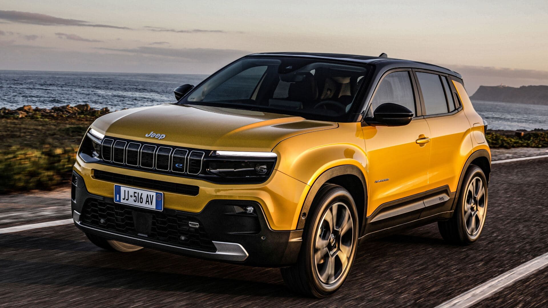 2025 Jeep Avenger in gold three quarter view driving on road with sea background 2025 Jeep Avenger in gold three quarter view driving on road with sea background