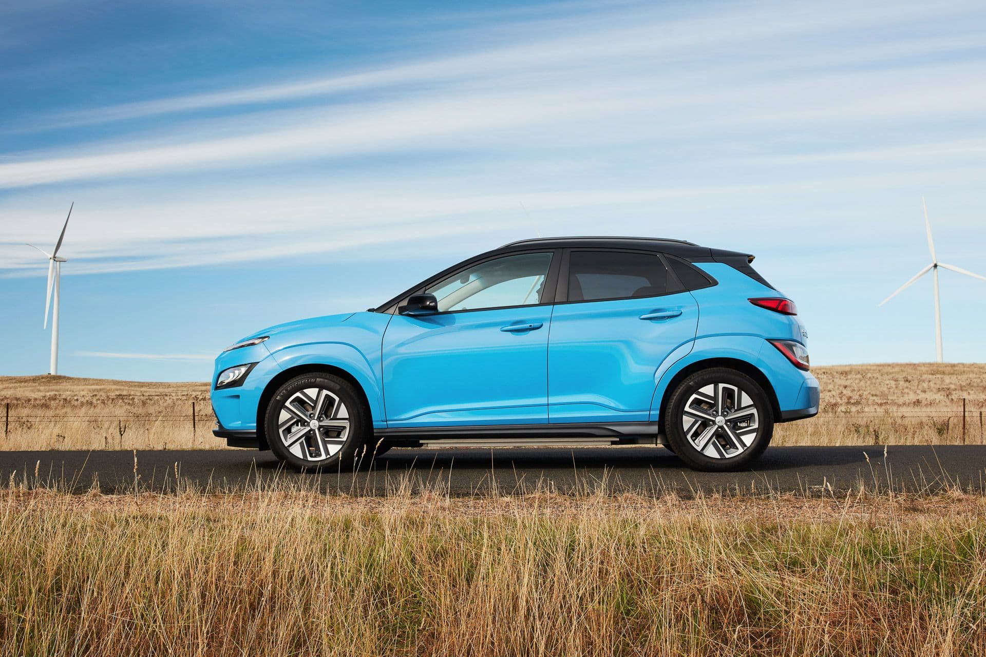 Blue with black roof Hyundai Kona Electric side view in front of wind turbines Blue with black roof Hyundai Kona Electric side view in front of wind turbines