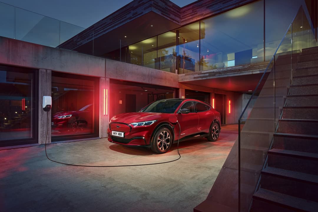 Red Ford Mustang Mach-E Premium charging from wall box in front of modern home Red Ford Mustang Mach-E Premium charging from wall box in front of modern home
