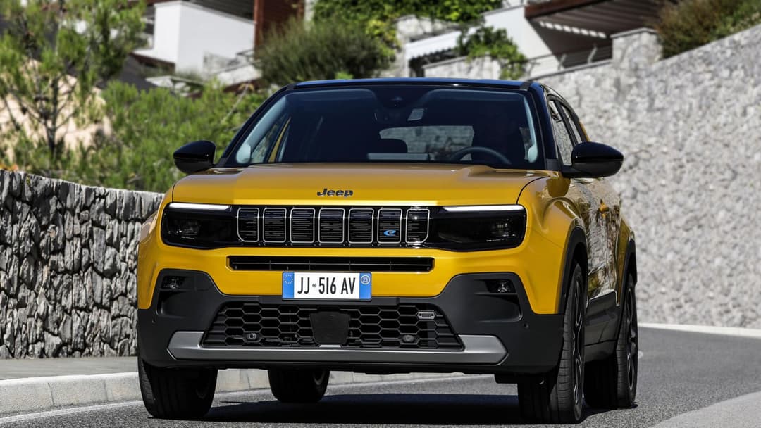 2025 Jeep Avenger in gold front view driving on road with stone wall background 2025 Jeep Avenger in gold front view driving on road with stone wall background