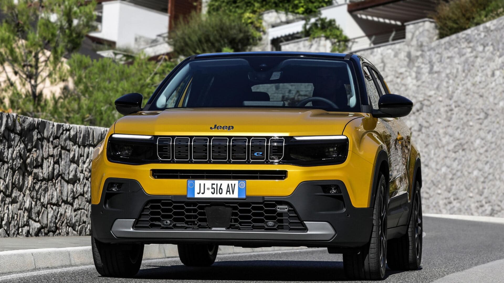 2025 Jeep Avenger in gold front view driving on road with stone wall background 2025 Jeep Avenger in gold front view driving on road with stone wall background