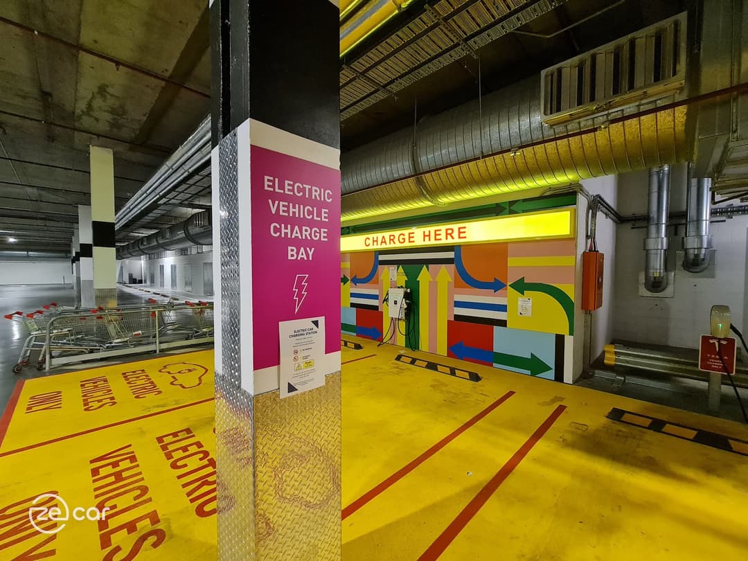 EV AC charger with yellow 'Charge Here' sign and pink 'Electric Vehicle Charge Bay' sign EV AC charger with yellow 'Charge Here' sign and pink 'Electric Vehicle Charge Bay' sign