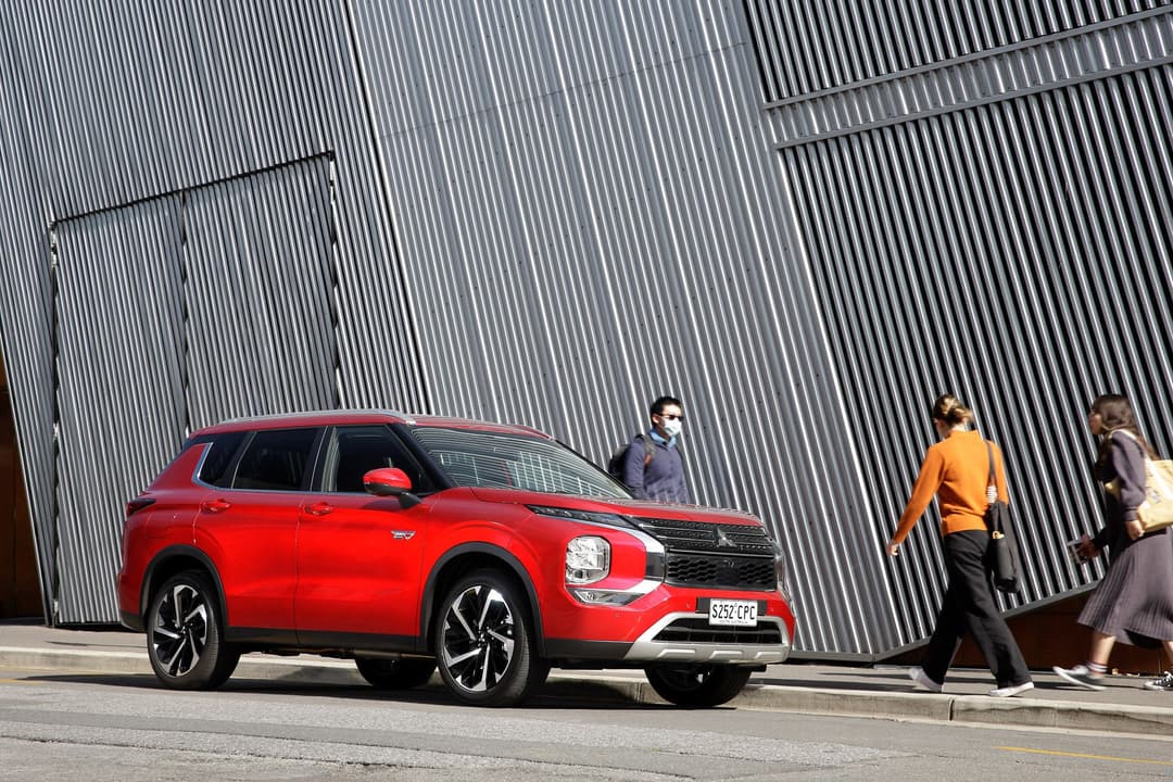 Pedestrians walk beside red Mitsubishi Outlander PHEV in city Pedestrians walk beside red Mitsubishi Outlander PHEV in city