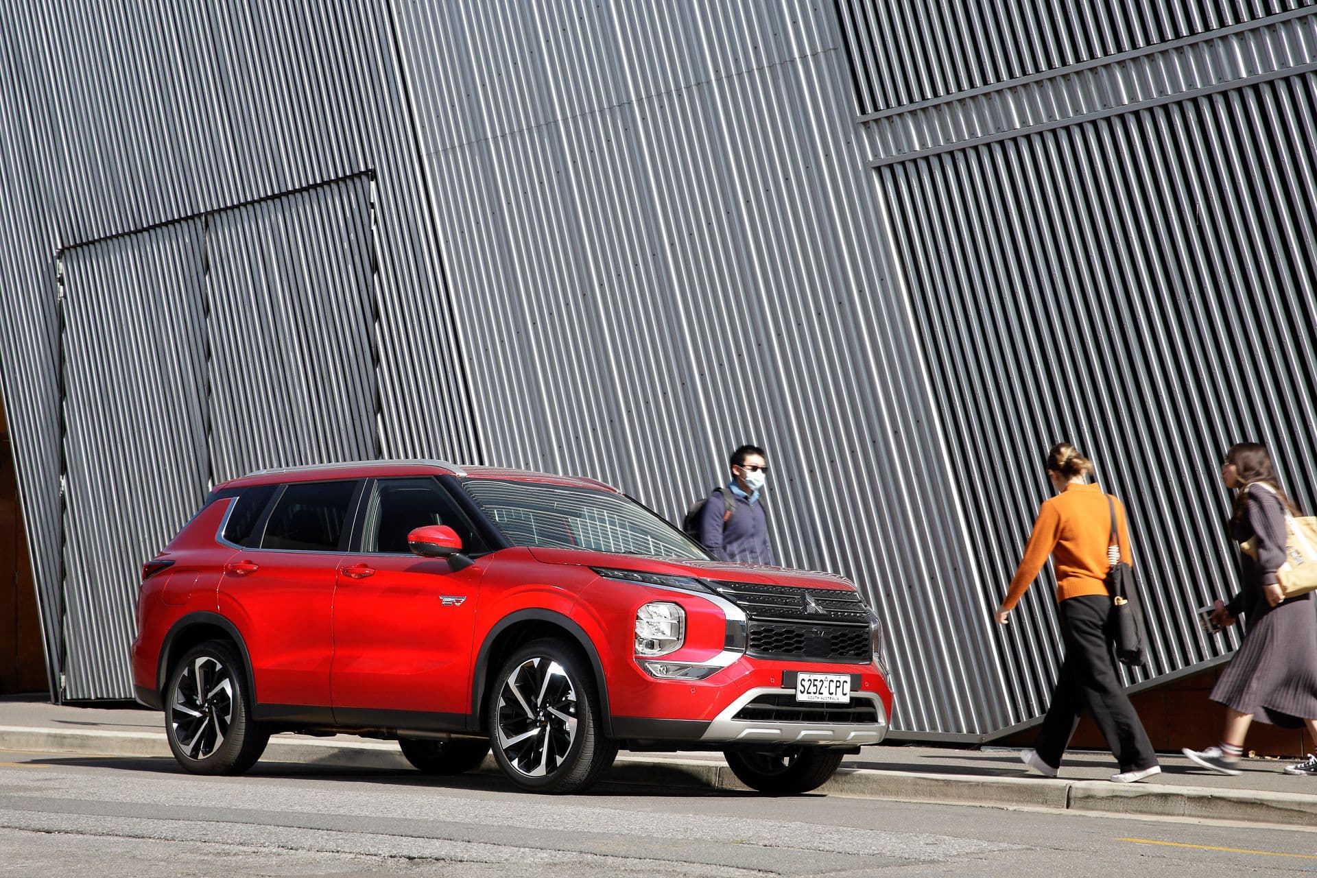 Pedestrians walk beside red Mitsubishi Outlander PHEV in city Pedestrians walk beside red Mitsubishi Outlander PHEV in city
