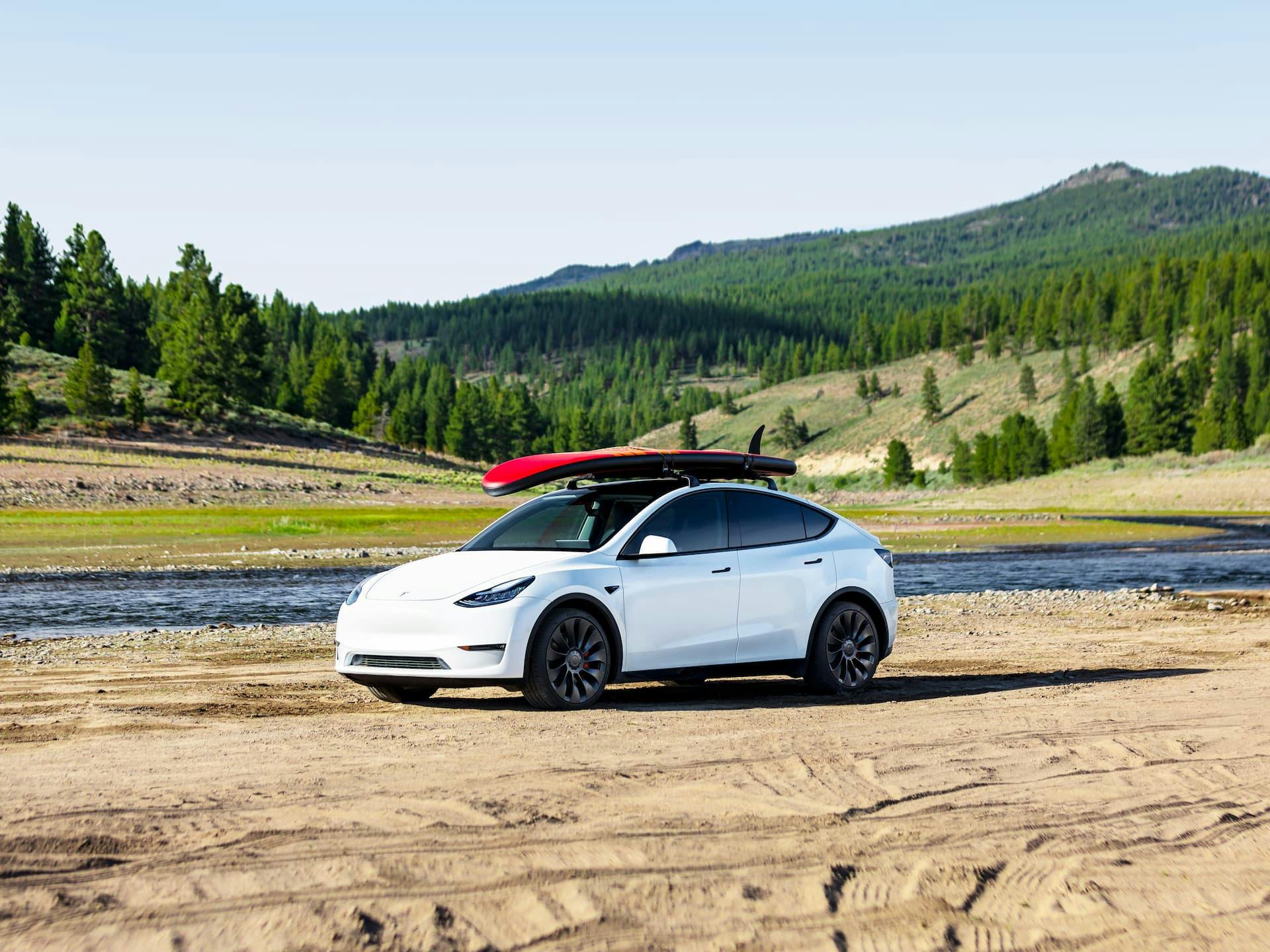 White Tesla Model Y with surfboard on roof beside lake